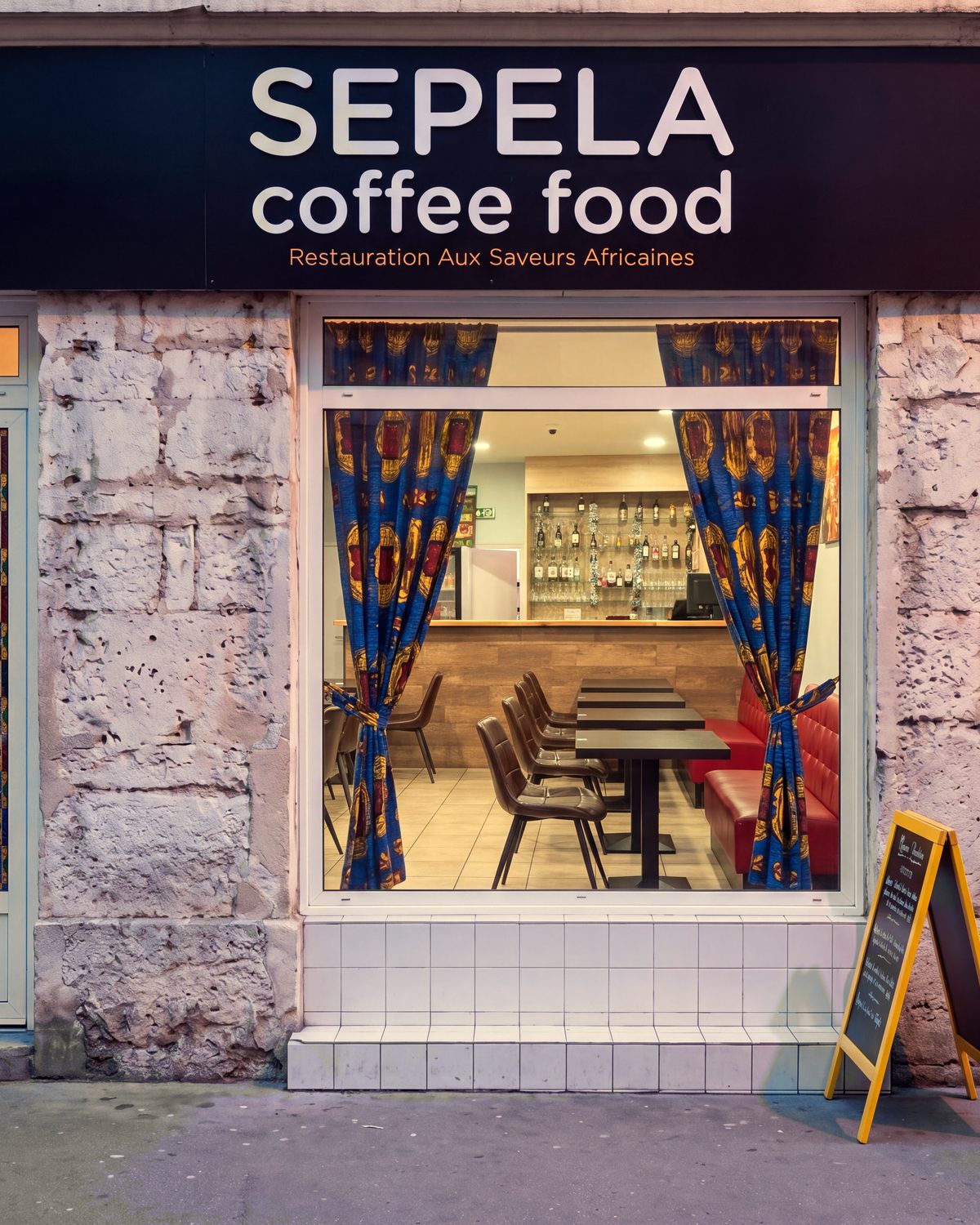 Façade du restaurant africain Sepela au 107 Grande Rue de la Guillotière, Lyon 7, la salle éclairée derrière la vitrine aux rideaux wax